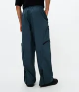Pantalón de corte holgado en color azul petróleo, confeccionado en microfibra de poliéster. Presenta un diseño utilitario con múltiples bolsillos funcionales, cierres visibles y detalles técnicos en las piernas.