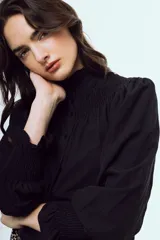Camisa negra de manga larga con cuello alto gorguera y detalles fruncidos con nido de abeja en el pecho y los puños. Cierre frontal con botones.