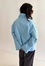 Campera celeste de algodón con diseño versátil de cuello alto envolvente, cierre frontal cruzado con botones metálicos a presión y corte boxy.