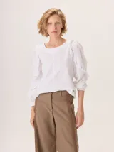 Camisa blanca de algodón con cuello redondo y mangas largas abullonadas con detalle de volados en los hombros y puños elásticos.