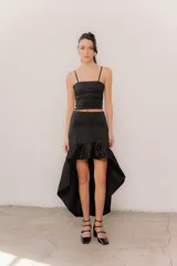 Pollera midi de tafeta color negro, con diseño asimétrico de corte mullet (más corta adelante y larga atrás), tiro alto y detalle de volado fruncido en el ruedo.