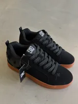 Championes DC Court Graffik color negro con suela de goma marrón. Presentan cordones gruesos negros, logo de DC en la lengüeta y en el lateral, y detalles de costuras.