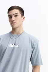 Remera de manga corta color celeste claro, con cuello redondo y estampado gráfico grande en la espalda con la palabra "BLESSED" y frases como "BE GRATEFUL EVERY MOMENT". Incluye el logo de la marca Mistral.