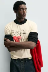 Camiseta de corte regular, color crema, con cuello redondo y ribetes en las mangas y cuello en color negro. Presenta un estampado frontal con un corazón rojo y texto en negro que dice "Everything beats for something".