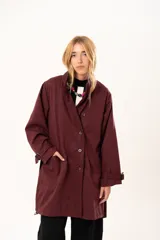 Trench de corte holgado color bordó, con cuello camisero, cierre frontal con botones y cinturón ajustable con hebilla. Presenta mangas largas con detalles de correas en los puños y bolsillos laterales.