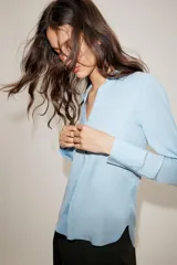Camisa de cuello solapa y escote en pico, con manga larga y cierre frontal mediante botones. Presenta un diseño fluido con bajo asimétrico y aberturas laterales.
