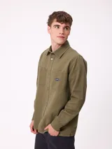 Camisa de gabardina color verde oliva, con cuello clásico, cierre frontal de botones y dos bolsillos frontales con parche. Incluye un pequeño parche con el logo de la marca en el bolsillo izquierdo.