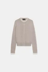 Chaqueta de punto color beige, de manga larga y corte corto, con cuello redondo y puños terminados en puntilla blanca a contraste.