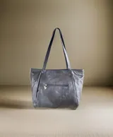 Cartera de cuero color negro, de diseño amplio con asas largas para llevar al hombro. Presenta un detalle de pintura abstracta en tonos nude y marrones en la parte inferior, y un charm metálico colgante.