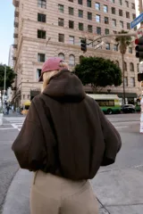 Chaqueta estilo bomber de corte oversize en color marrón oscuro, confeccionada en tejido suave. Presenta capucha con cordones ajustables, cierre frontal con cremallera, mangas largas con puños elásticos y bolsillos delanteros de vivo. El bajo cuenta con detalle de pliegues.