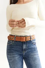 Cinto de cuero color suela con diseño de tachas metálicas circulares en tonos plateados y dorados. Cuenta con hebilla metálica rectangular de estilo clásico.