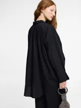 Camisa negra de corte oversize y manga larga con puños abotonados. Presenta un cuello clásico con un lazo fino para anudar en la parte superior y un diseño con bajo asimétrico, siendo más larga en la parte posterior.