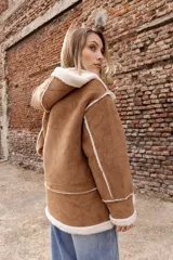 Campera estilo gamulán color tostado con interior y bordes de corderito sintético color crema. Presenta capucha amplia, cierre frontal con trabas y broches metálicos, y bolsillos delanteros grandes. Posee un calce relajado y diseño de textura suave.