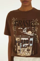 Remera marrón de manga corta y cuello redondo, con estampa frontal temática de Brasil que incluye palmeras, arquitectura colonial y aves.