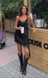 Vestido corto de satén negro con escote en V y breteles finos. Presenta detalles de encaje en el escote y en el ruedo asimétrico, con una caída fluida y elegante.
