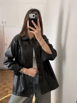 Sobrecamisa oversize de cuero sintético negro, con solapa clásica, cierre frontal con botones y flecos decorativos en el pecho. Cuenta con mangas largas y bolsillos laterales.