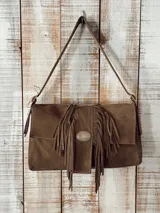 Cartera tipo bandolera de cuero gamuzado color marrón, con solapa frontal decorada con flecos y una hebilla metálica ovalada grabada. Cuenta con una correa larga ajustable para el hombro.