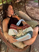 Bolso tejido a mano con hilo de trapillo, eco-cuero y textiles, con un diseño central de querubín coronado y alas, sobre un fondo de franjas rojas, blancas y azules. Posee asas cortas tejidas en color celeste claro.