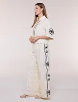 Conjunto de dos piezas negro compuesto por camisa de manga corta con cuello camisero y pantalón palazzo de tiro alto. Presenta detalles de bordados florales en contraste a lo largo de los laterales del pantalón y en las mangas.