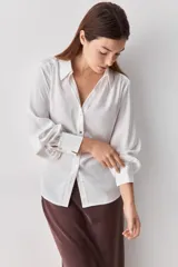 Camisa blanca de manga larga con textura suave y caída liviana, cuello camisero, puños anchos y fruncidos sutiles en los hombros.