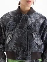 Chaqueta estilo bomber confeccionada en gabardina con estampado de damasco. Presenta cierre frontal oculto por carterón, bolsillos con tapa en forma de V y detalles tejidos en cuello y ruedo.