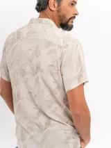 Camisa de manga corta color beige con estampado tropical de hojas, cuello camisero abierto y cierre frontal de botones.
