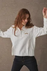 T-shirt de manga larga confeccionada en mezcla de algodón y lino, color blanco, con cuello camisero, cierre frontal parcial con cremallera, puños abotonados y aberturas laterales en el bajo. Cuenta con un pequeño bordado distintivo en el pecho.