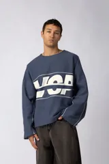 Buzo oversize de algodón rústico de alto gramaje, color azul grisáceo, con diseño boxy, cortes con bordes deshilachados y estampa frontal con letras blancas.