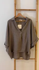Blusa holgada de lino color marrón topo, con cuello camisero y escote en V. Presenta mangas cortas con puños doblados y un ruedo asimétrico más largo en la espalda.