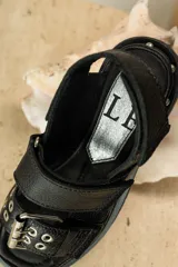 Sandalias negras de cuero con diseño de plataforma dentada, tiras ajustables con hebillas metálicas y detalle de tachas plateadas en el lateral de la suela.