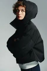 Anorak corto de color negro con acolchado interior, cuello subido y capucha. Presenta mangas largas con puños elásticos, bolsillos delanteros con cierre y bajo ajustable con cordones. Cierre frontal con cremallera termosellada y botones a presión.