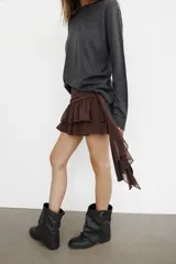 Falda pantalón de tiro bajo en color negro, con cintura drapeada y detalle de tira lateral colgante. Presenta un diseño con volantes en el bajo y cierre de cremallera metálica.