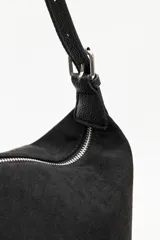 Cartera de gamuza negra con textura suave y acabado aterciopelado. Cuenta con una silueta amplia y flexible, cierre superior metálico, correa regulable y detalles laterales con hebillas.