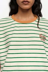 Remera estilo boxy de algodón con diseño de rayas horizontales verdes sobre fondo crudo. Cuenta con cuello redondo marrón tostado, mangas cortas caídas y detalle de logo bordado en el pecho.