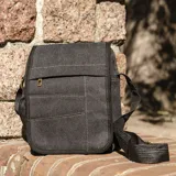 Morral bandolera de tela canvas color gris oscuro con detalles en negro. Tiene tapa con cierre, bolsillo frontal con cierre bajo la tapa, bolsillo trasero con cierre y correa larga regulable.
