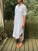 Vestido camisero largo de lino blanco, con cuello camisero, manga corta enrollada y cierre frontal de botones de nácar. Presenta aberturas laterales en el bajo.