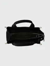 Cartera tipo tote bag de lona negra con letras blancas estampadas en el frente. Cuenta con doble asa superior, correa ajustable y desmontable, y detalles laterales en contraste.