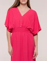 Vestido largo amarillo de corte fluido con escote en V, mangas cortas amplias y apliques de tachas en el escote y puños. Posee cintura elastizada fruncida y falda larga con tajos laterales en el ruedo.