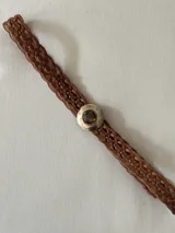 Cinto trenzado de cuero color camel con hebilla circular metálica grabada y detalle de piedra central.