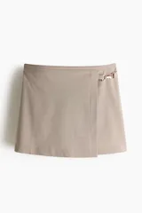 Pollera mini de corte cruzado con diseño a cuadros en tonos beige y marrón. Presenta una hebilla metálica decorativa en el lateral y cierre oculto en la parte posterior.