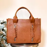 Cartera de cuero color tostado, estructurada, con doble asa de mano, correa desmontable y dos bolsillos frontales verticales con cierre. Presenta detalles de costuras pespunteadas y el logo de la marca grabado en la parte inferior.