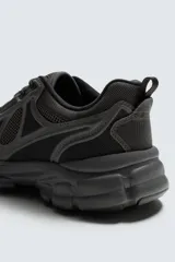 Zapatilla deportiva de estilo running, completamente negra, con capellada de malla y superposiciones sintéticas. Presenta una suela gruesa y voluminosa con diseño segmentado.