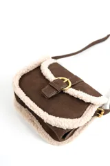 Cartera bandolera marrón de gamuza sintética con detalles de corderito sintético color beige en los bordes. Cuenta con una solapa frontal con cierre de hebilla metálica dorada y correa ajustable.