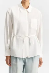 Camisa blanca de manga larga con cuello camisero, bolsillo frontal superior y tiritas ajustables en los costados para atar en la cintura.