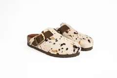 Zueco con capellada de pelo de vaca con manchas marrones y negras sobre base blanca, plantilla anatómica de cuero y hebilla lateral ajustable.