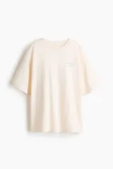 Remera oversized de punto suave de algodón color beige, con cuello redondo acanalado, hombros caídos y diseño estampado en el pecho con la inscripción "Taormina Sicily" en color azul.