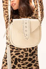 Cartera de cuero con textura efecto croco en color off-white. Presenta una solapa frontal decorada con tachas metálicas, un colgante de flecos largo y una correa ajustable. Incluye un bolsillo trasero con cierre y herrajes metálicos.