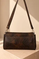 Cartera de cuero vacuno color marrón oscuro, con diseño trenzado y solapa frontal con placa metálica dorada grabada con la marca PECARÍ. Cuenta con correa larga para hombro y herrajes metálicos plateados.