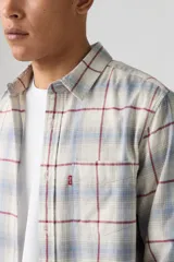 Camisa de manga larga con diseño a cuadros en tonos beige, azul y rojo. Presenta cuello clásico, cierre frontal con botones y un bolsillo en el pecho con etiqueta roja distintiva.