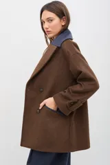 Tapado de paño de lana color marrón con silueta oversize y hombros caídos. Presenta doble abotonadura frontal, solapas amplias con detalle en contraste azul, y mangas con ajuste en los puños.